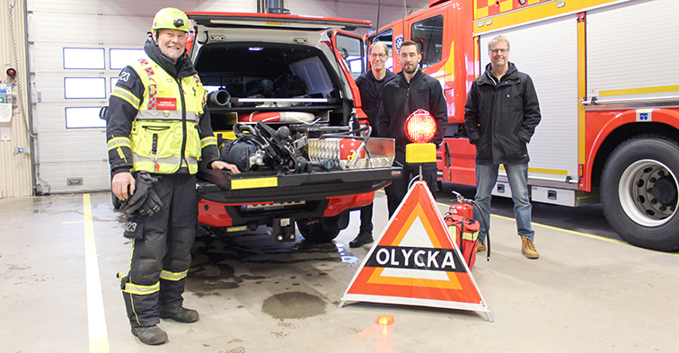 En man i larmkläder står vid sidan av en brandbil med bakluckan uppe. Där finns olika typer av verktyg och utrustning. Vid andra sidan står tre män i svarta kläder.