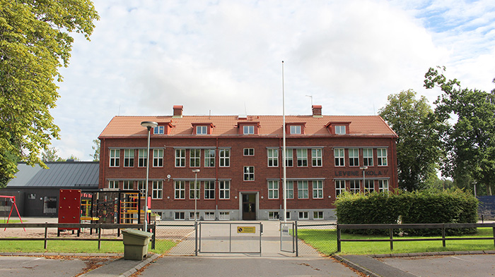 Röd skolbyggnad med tre plan, skolgård framför.